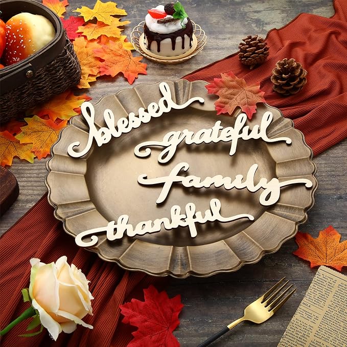 A metallic plate holding four wooden cutouts that read blessed, grateful, family, and thankful, surrounded by autumn leaves, pinecones, a small cake, and a fork on a wooden table.