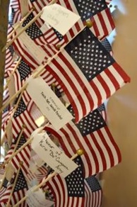 A series of small American flags on sticks, each featuring a hand-written memorial tag attached to the flagpole.