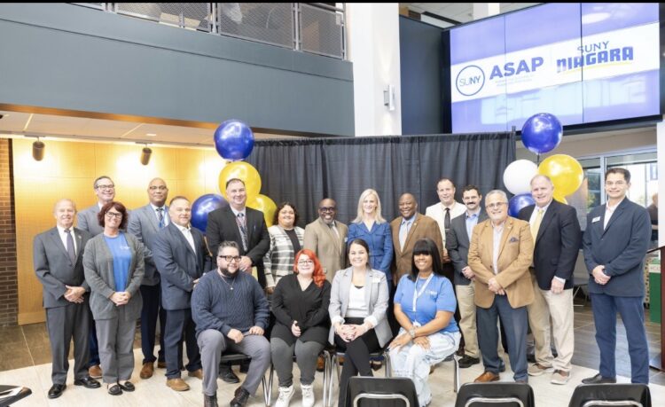 A group of 17 people stand and sit together for a photo in front of a black curtain and a large screen displaying the SUNY Niagara ASAP logo.