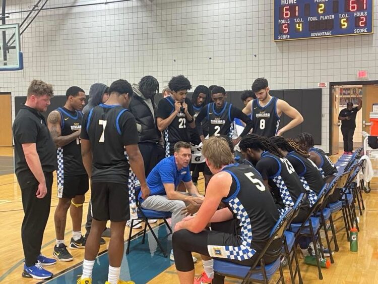 A basketball coach sits on a chair to talk to his team during a huddle on the court.