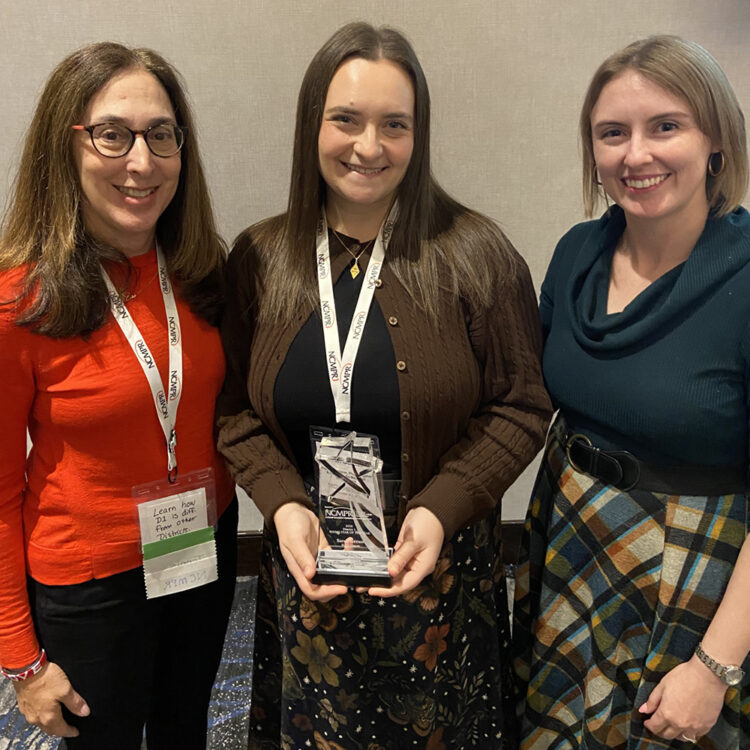 Three women stand together, with the center woman holding an award trophy and all wearing conference lanyards.