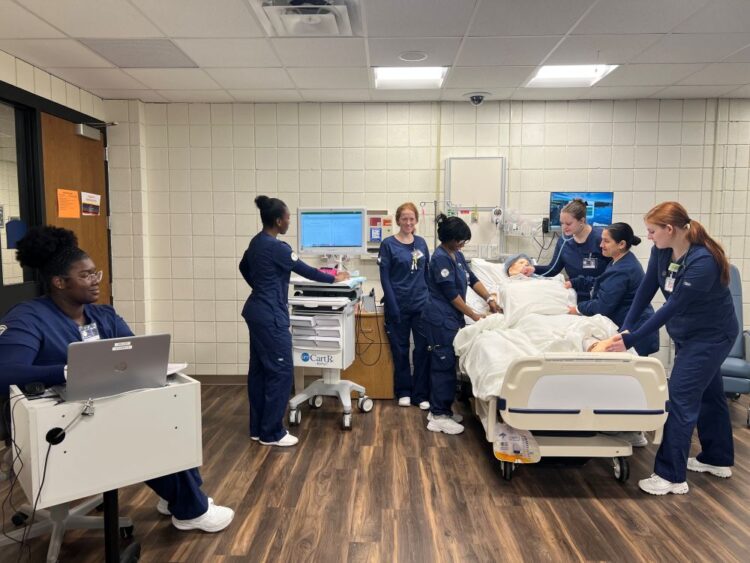 Nursing students in scrubs practice patient care around a medical mannequin in a hospital bed, with one student monitoring a laptop nearby.