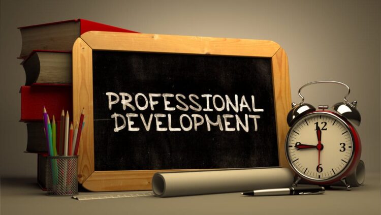 A chalkboard with the words Professional Development written on it, positioned next to a stack of books, a pencil holder, a rolled paper, and an alarm clock.