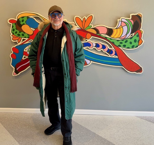 A man in a green coat, baseball cap, and blue-rimmed glasses stands in front of a colorful, abstract wall-mounted sculpture.